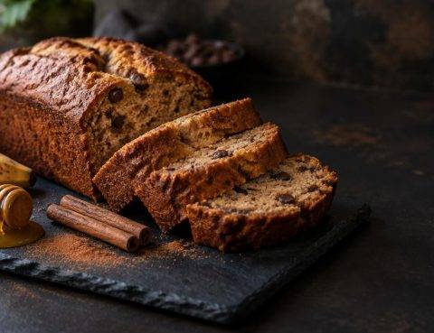 Σπιτικό banana bread σε φέτες πάνω σε πλάκα με μπανάνα, μέλι και ξυλάκια κανέλας